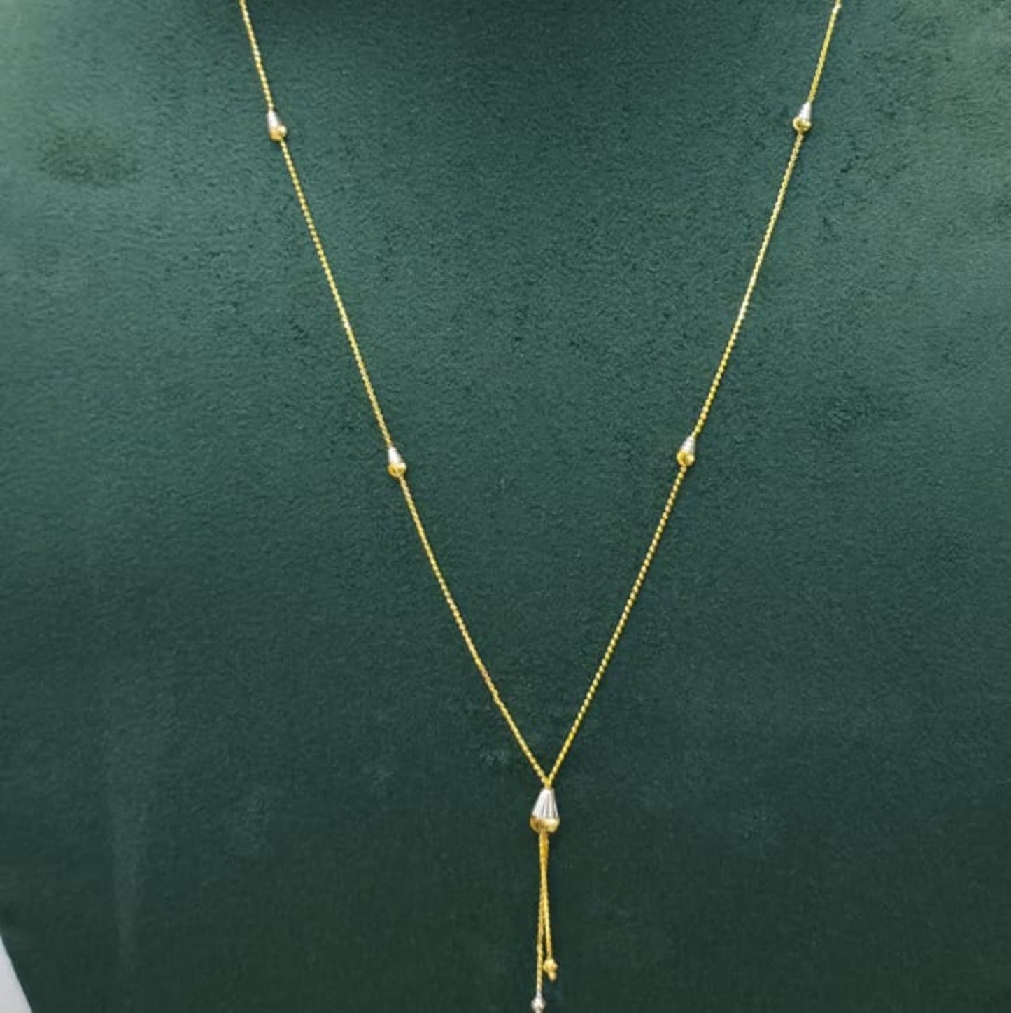 A delicate gold chain necklace with small bead accents and a teardrop-shaped pendant, displayed on a dark green velvet jewelry bust against a light gray background.