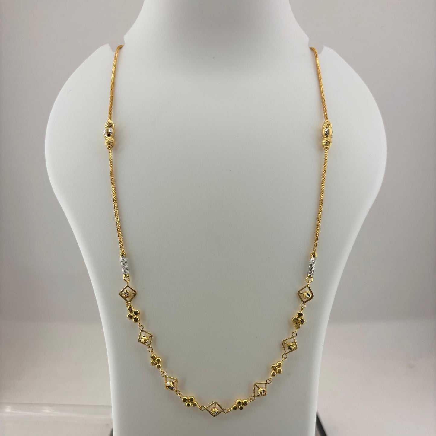 A gold chain necklace with small diamond-shaped and floral motifs displayed on a white mannequin bust, with a digital scale visible underneath.