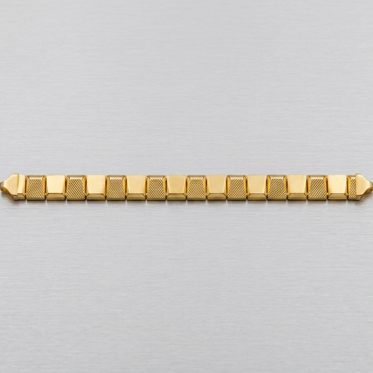 A gold-toned metal link bracelet with a textured pattern on the links, displayed horizontally on a brushed silver metal surface.