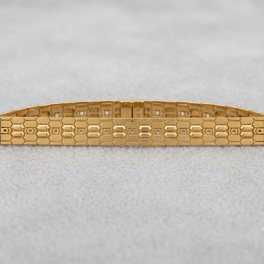 A gold-tone metal bracelet with a textured honeycomb pattern and small clear stones set along the top, displayed on a light gray textured background.