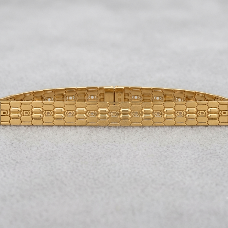 A gold-tone metal bracelet with a textured honeycomb pattern and small clear stones set along the top, displayed on a light gray textured background.