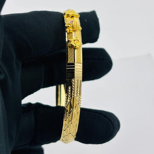 A gold-plated bangle bracelet with detailed engravings and a decorative clasp, held by a hand wearing a black glove against a plain light gray background.