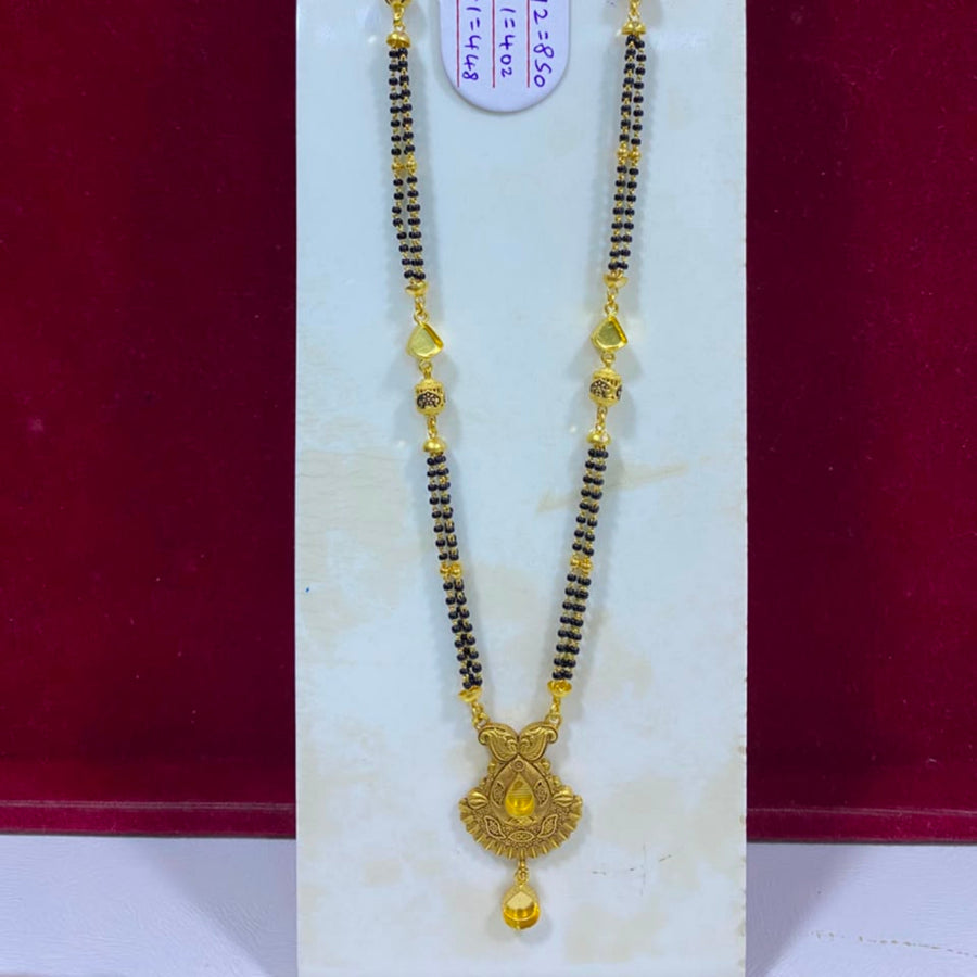 A traditional Indian gold and black beaded necklace (Murugai) with a detailed gold pendant, displayed on a white jewelry stand against a dark red background.