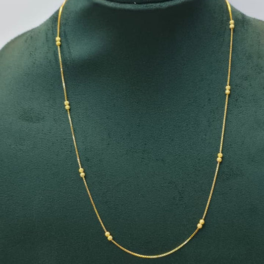 A delicate gold chain necklace with small bead-like spaced accents, displayed on a dark green velvet jewelry bust against a light gray background.