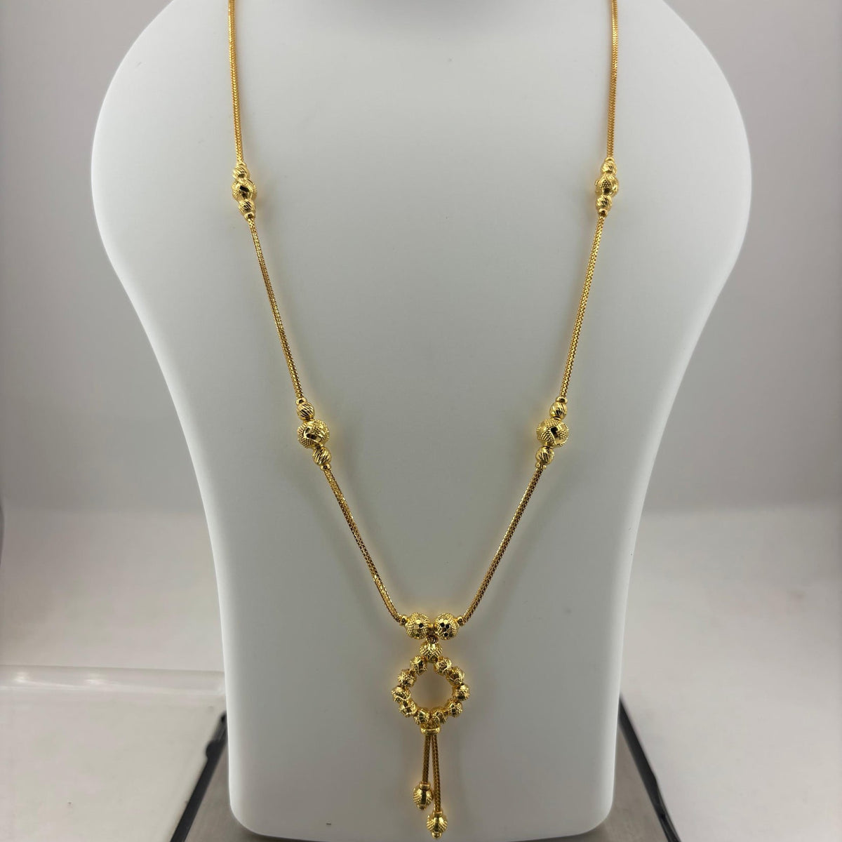 A gold necklace displayed on a white jewelry bust, with a digital scale visible underneath. The necklace features a delicate chain with small bead details and a central key-shaped pendant.