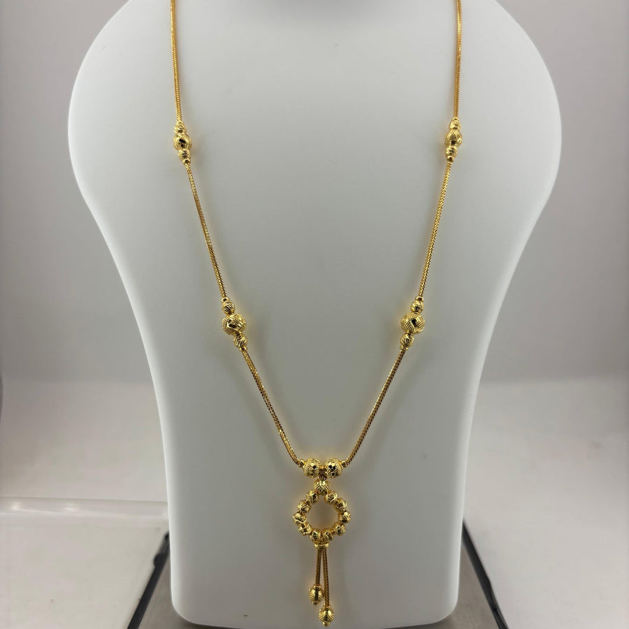 A gold necklace displayed on a white jewelry bust, with a digital scale visible underneath. The necklace features a delicate chain with small bead details and a central key-shaped pendant.