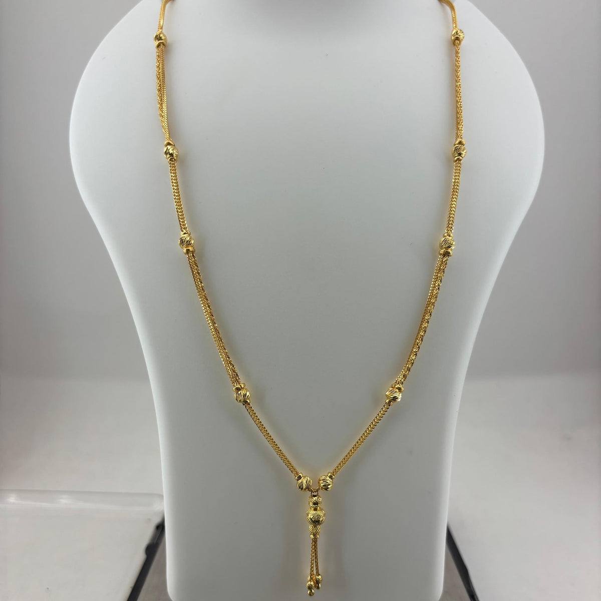 A gold necklace displayed on a white jewelry bust, with a digital scale visible underneath. The necklace features a delicate chain with small bead-like links and a central pendant with a tassel drop.