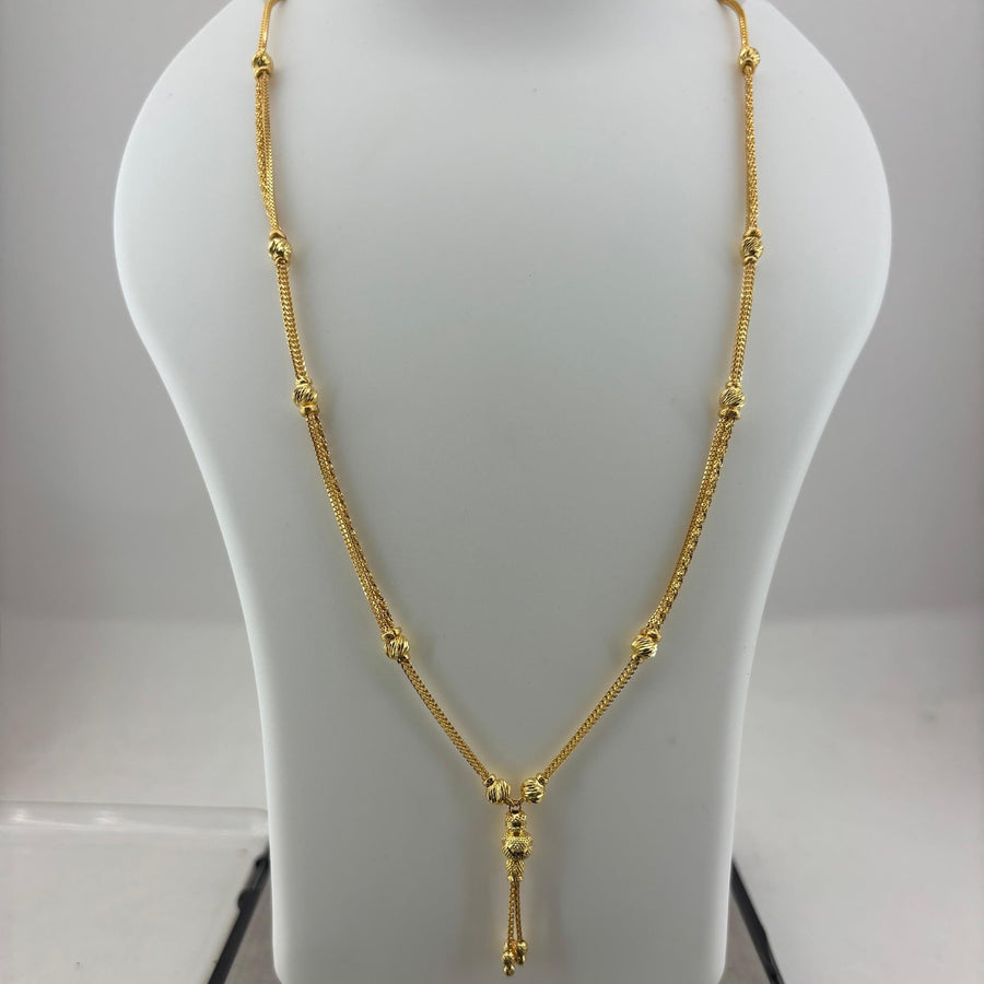A gold necklace displayed on a white jewelry bust, with a digital scale visible underneath. The necklace features a delicate chain with small bead-like links and a central pendant with a tassel drop.