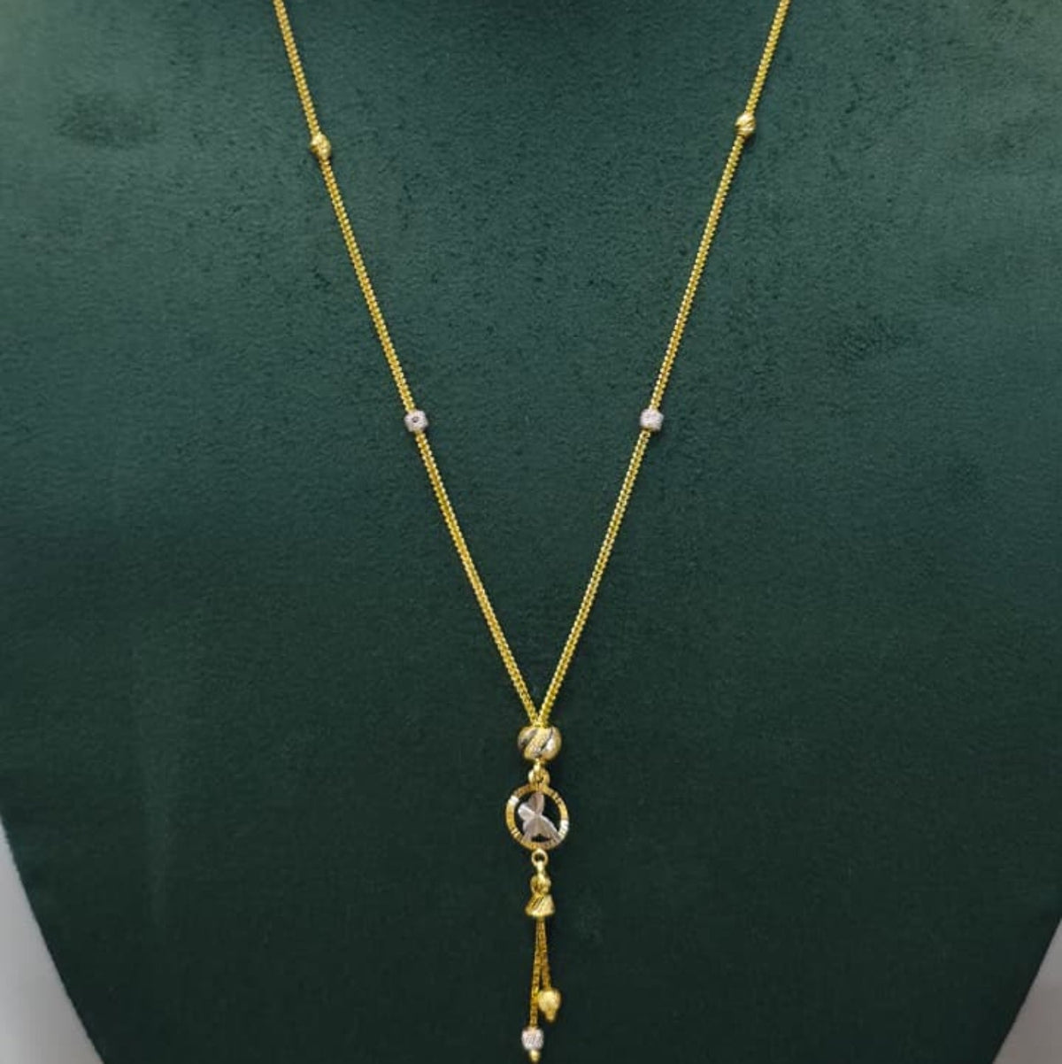 A gold chain necklace with a circular pendant and dangling tassels, displayed on a dark green velvet jewelry bust against a light gray background.