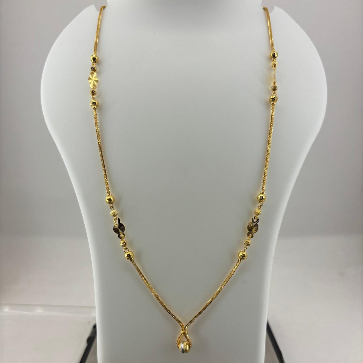 A gold necklace with small bead details and a teardrop pendant, displayed on a white jewelry bust with a digital scale underneath.