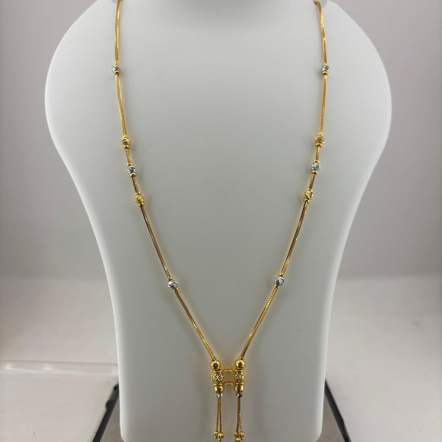 A gold necklace displayed on a white mannequin bust, with a digital scale visible underneath. The necklace features a Y-shaped design with small silver-toned bead accents and a central gold pendant with dangling chains.