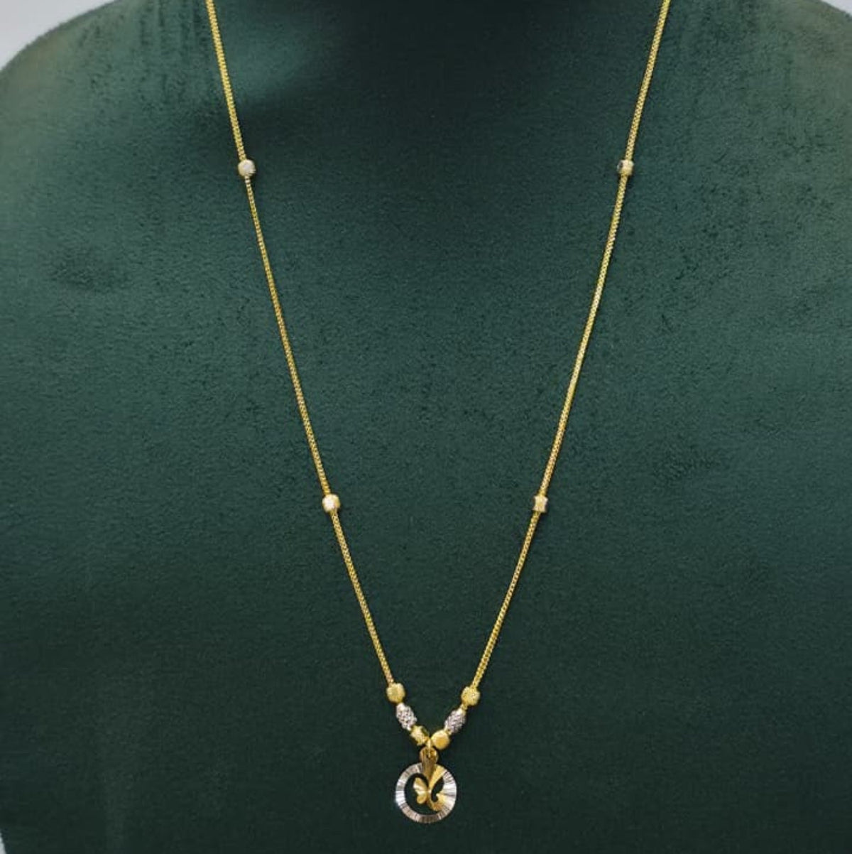 A delicate gold chain necklace with small bead accents and a round pendant, displayed on a dark green jewelry bust against a light gray background.