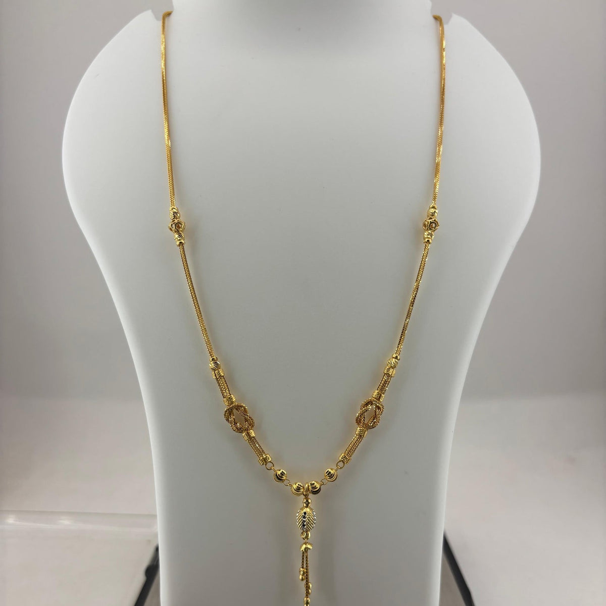 A gold necklace displayed on a white jewelry bust, with a digital scale visible underneath. The necklace features a textured chain with small circular and oval links and a dangling pendant.