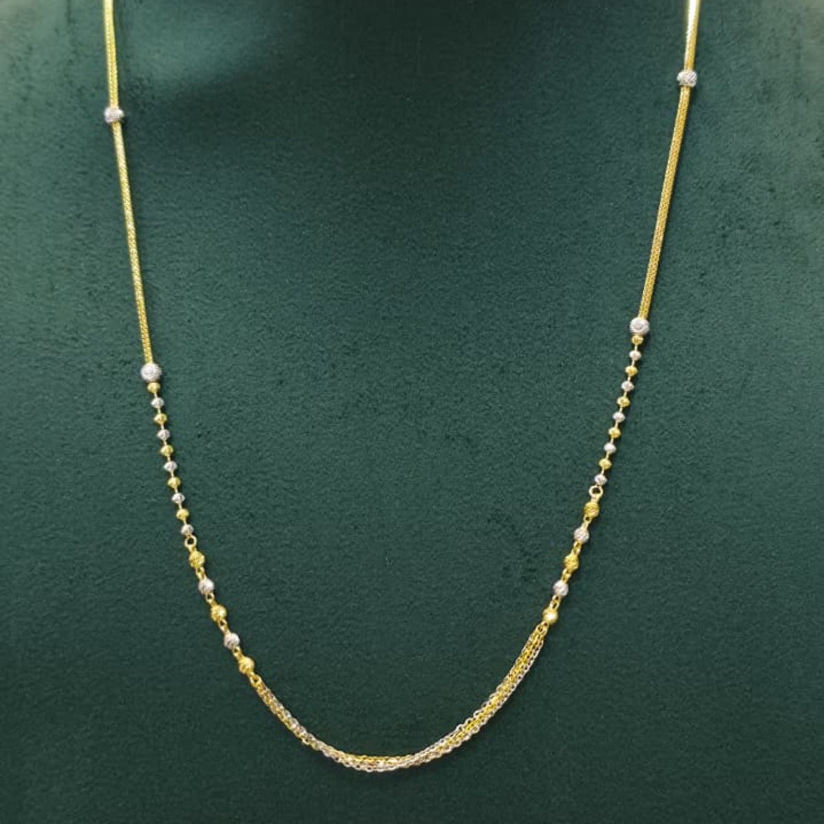 A delicate gold chain necklace with small silver bead accents, displayed on a dark green jewelry bust.