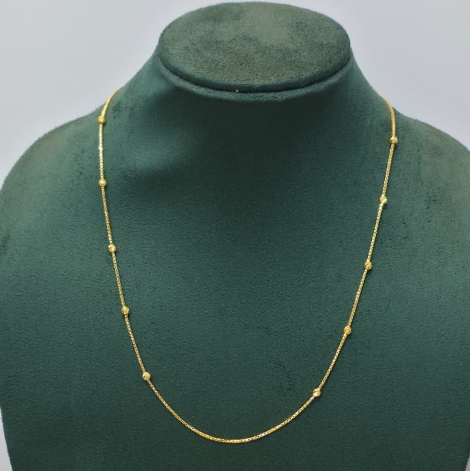 A delicate gold chain necklace with small spaced beads, displayed on a dark green velvet jewelry bust against a light gray background.