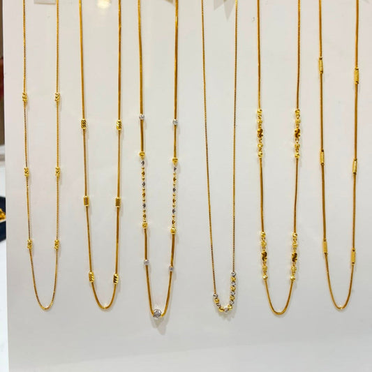Nine gold-tone chain necklaces with various bead and spacer details are displayed on a white vertical rack with price tags attached, set against a softly blurred indoor background.