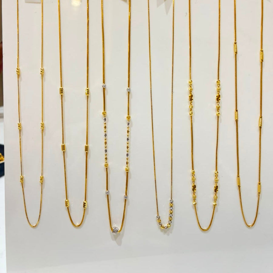 Nine gold-tone chain necklaces with various bead and spacer details are displayed on a white vertical rack with price tags attached, set against a softly blurred indoor background.