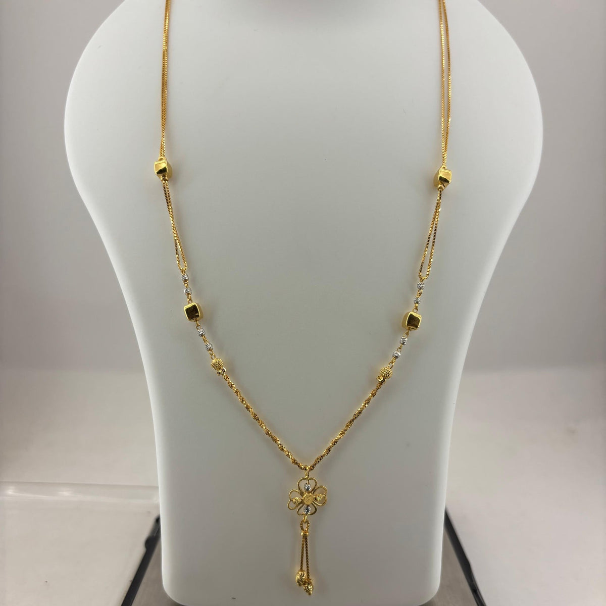 A gold necklace with a small floral pendant and dangling tassel, displayed on a white mannequin bust with a digital scale underneath.