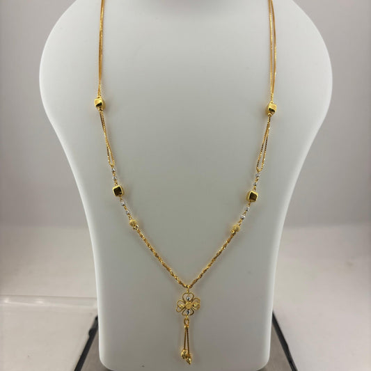 A gold necklace with a small floral pendant and dangling tassel, displayed on a white mannequin bust with a digital scale underneath.