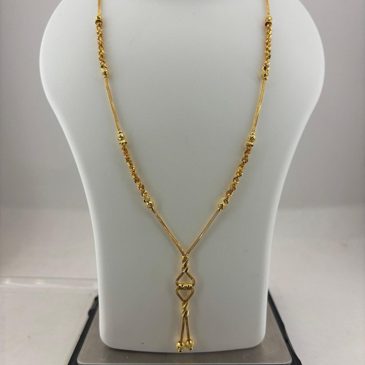 A gold necklace displayed on a white jewelry bust, with a digital scale underneath to show weight. The necklace features a textured chain and a pendant with a small central heart-shaped detail.