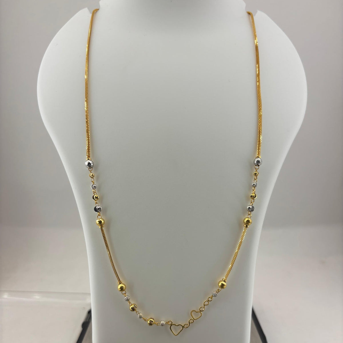 A gold necklace with small silver and gold beads and a heart-shaped clasp, displayed on a white jewelry bust sitting on a digital scale.