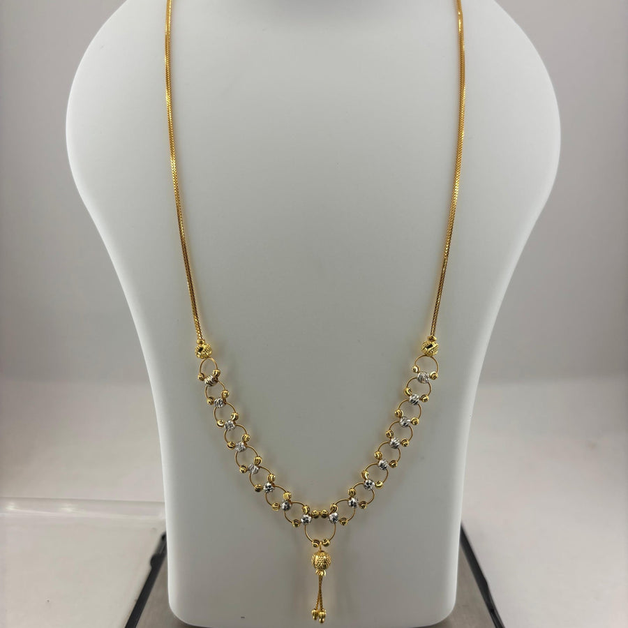 A gold necklace displayed on a white mannequin bust, with a digital scale visible underneath. The necklace features a delicate chain with a V-shaped arrangement of small gold links and a central pendant.