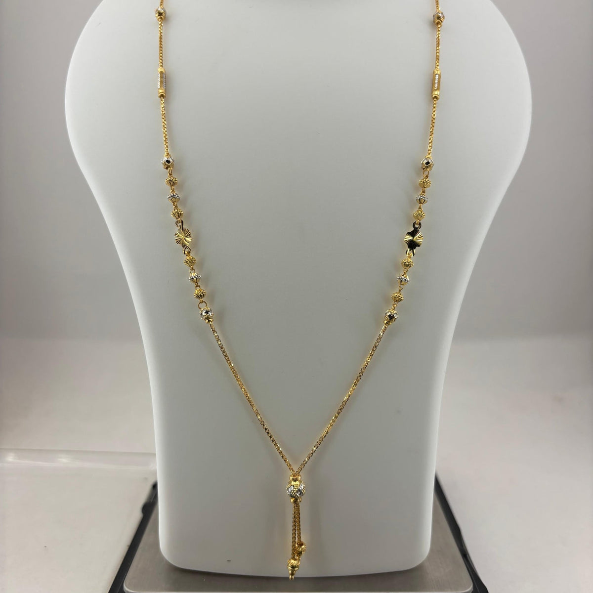 A gold necklace displayed on a white mannequin bust, with a digital scale visible underneath to show the weight. The necklace features a delicate chain with small bead details and a central pendant.