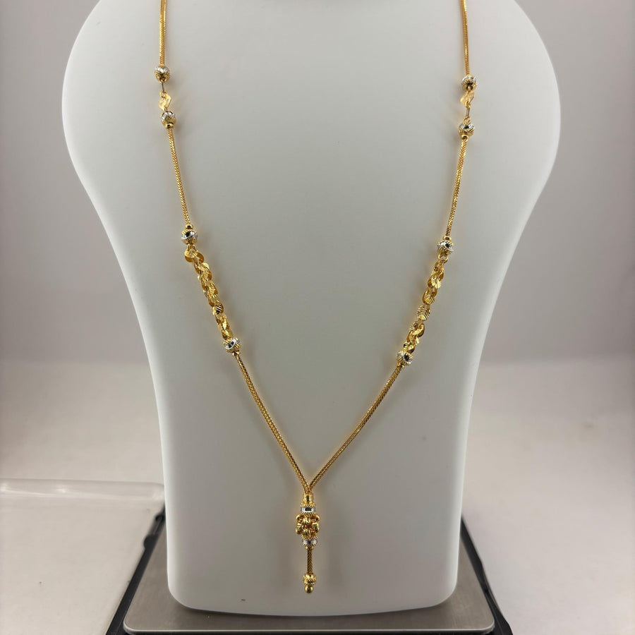 A gold necklace displayed on a white mannequin bust, with a digital scale visible underneath. The necklace features a delicate chain with textured bead clusters and a small key-shaped pendant.