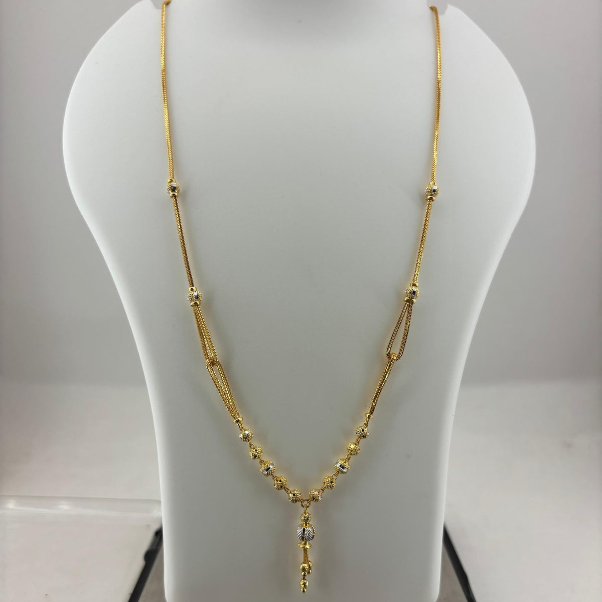 A gold necklace displayed on a white jewelry bust, with a digital scale visible underneath. The necklace features a delicate chain with small bead details and a central pendant with a clear stone.