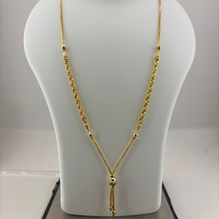 A gold necklace displayed on a white mannequin bust, with a digital scale visible underneath. The necklace features a twisted rope chain, small bead details, and a central pendant with hanging tassels.