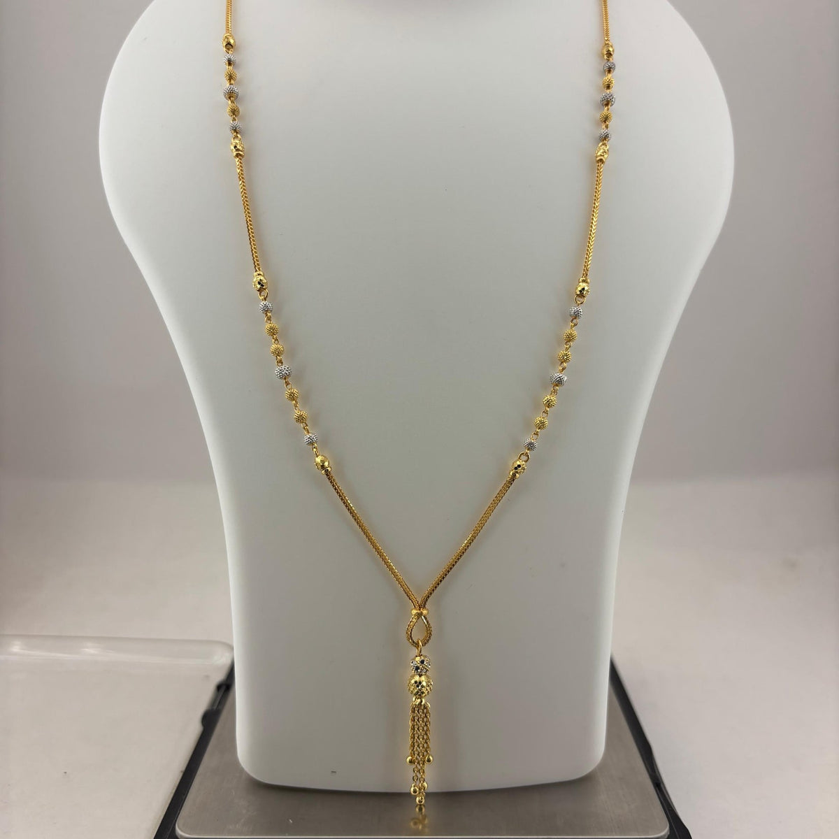 A gold necklace displayed on a white mannequin bust, with a digital scale visible underneath. The necklace features a delicate chain with small bead-like details and a textured pendant.