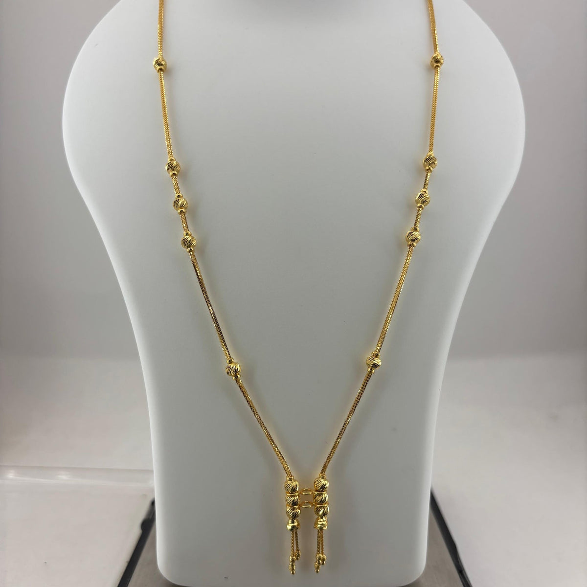 A gold necklace displayed on a white mannequin bust, with a digital scale visible underneath. The necklace features a delicate chain with small bead-like accents and a central pendant with three dangling chain ends.