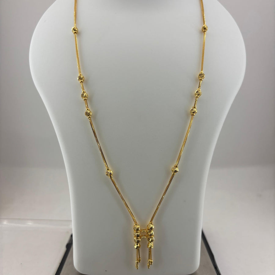 A gold necklace displayed on a white mannequin bust, with a digital scale visible underneath. The necklace features a delicate chain with small bead-like accents and a central pendant with three dangling chain ends.