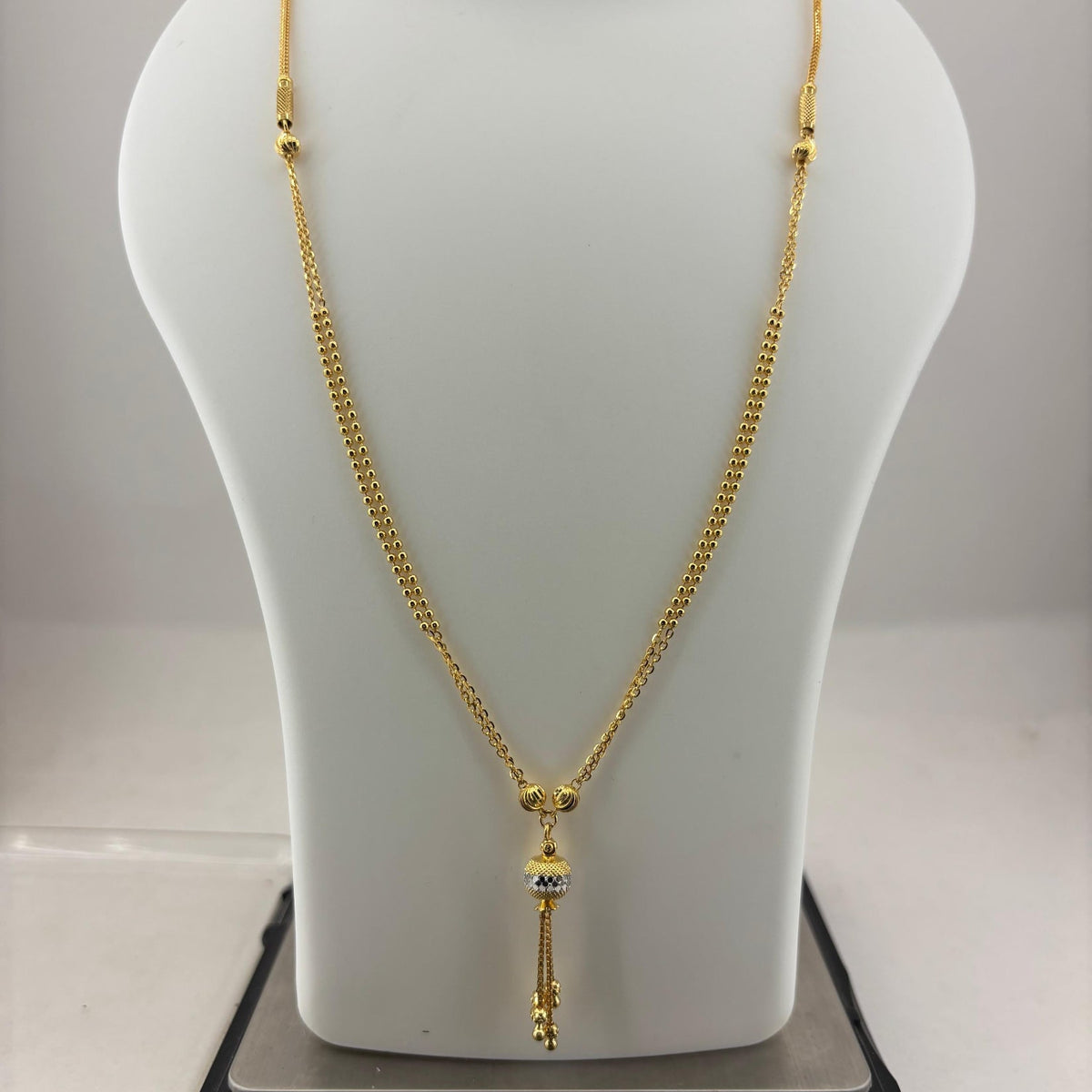 A gold-toned chain necklace with a decorative pendant is displayed on a white mannequin bust, with a digital scale visible underneath.