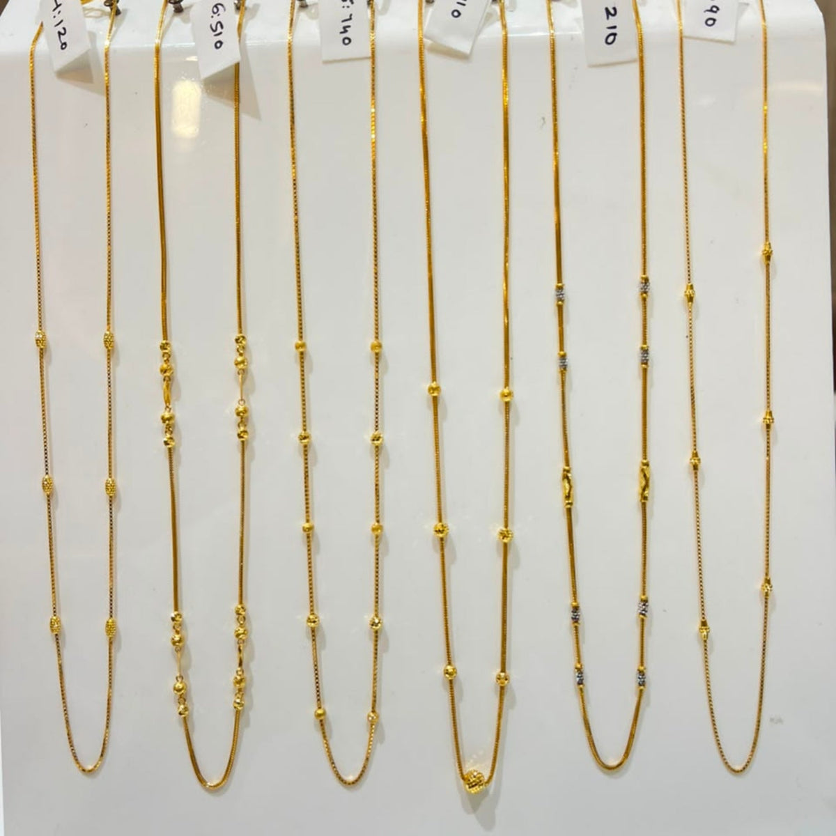 Nine gold beaded chains of varying lengths displayed on a white stand, each with a small white price tag attached.