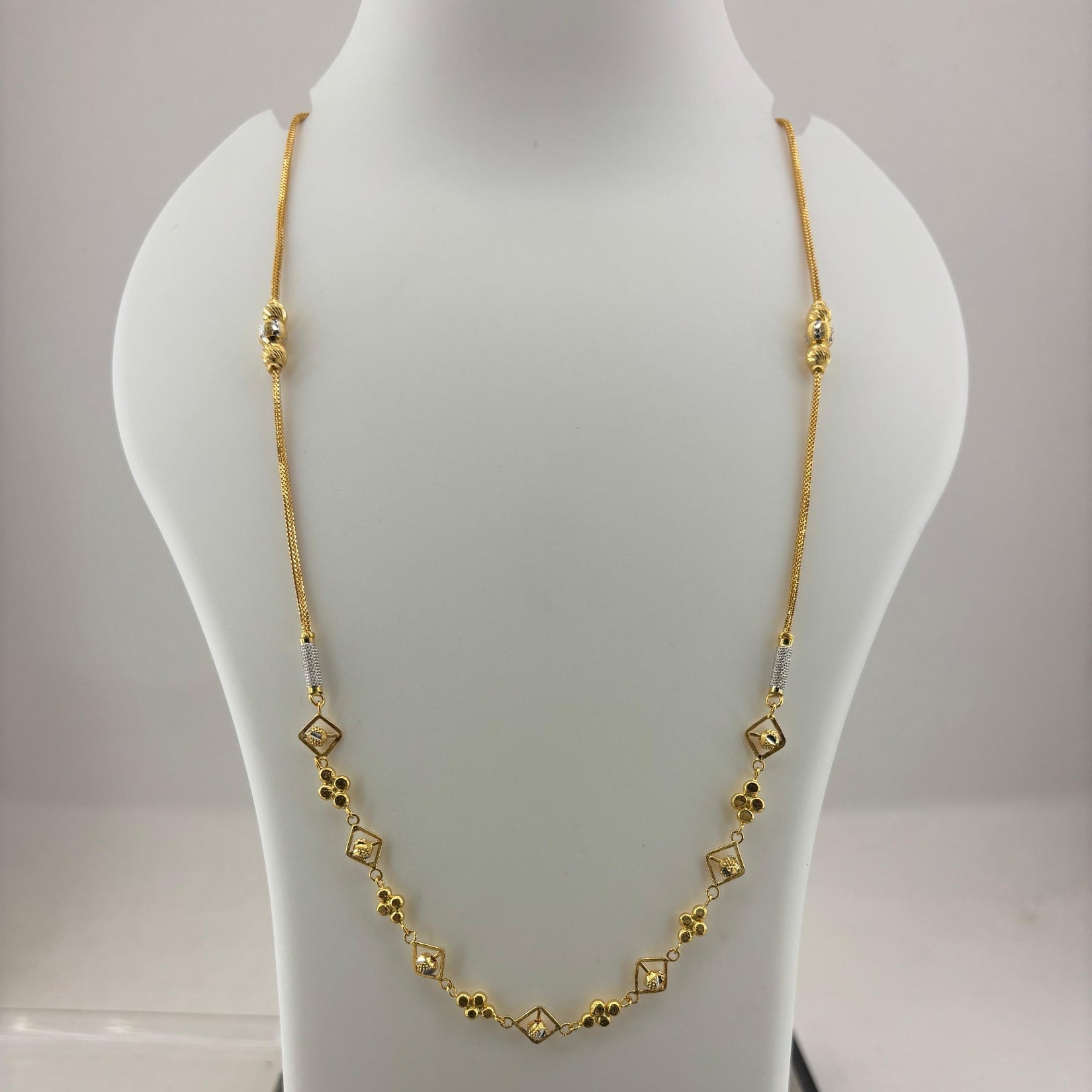 A gold chain necklace with small diamond-shaped and floral motifs displayed on a white mannequin bust, with a digital scale visible underneath.