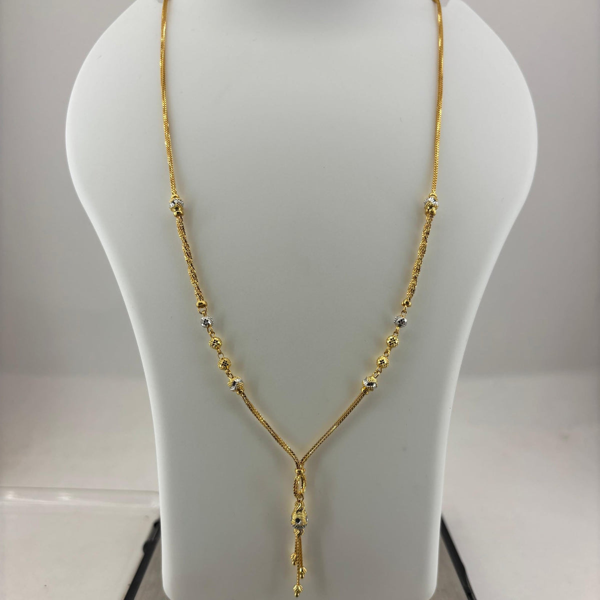 A gold necklace displayed on a white jewelry bust, with a digital scale visible underneath. The necklace has a Y-shaped design with small bead details along the chain.