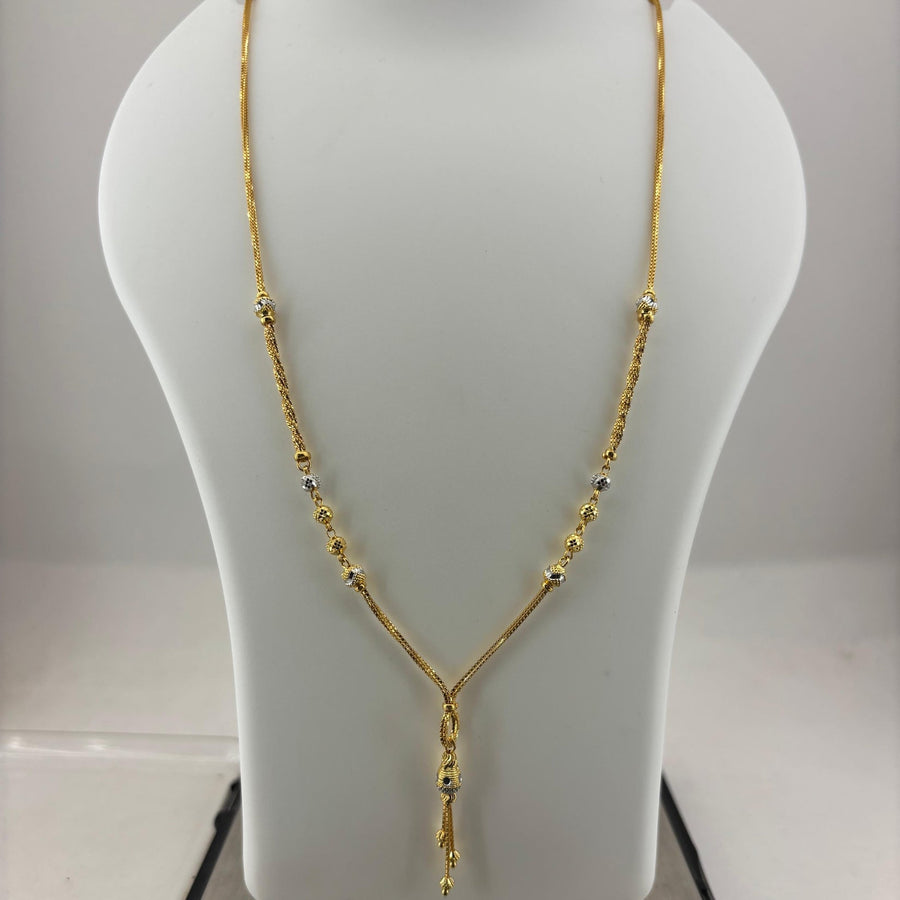 A gold necklace displayed on a white jewelry bust, with a digital scale visible underneath. The necklace has a Y-shaped design with small bead details along the chain.