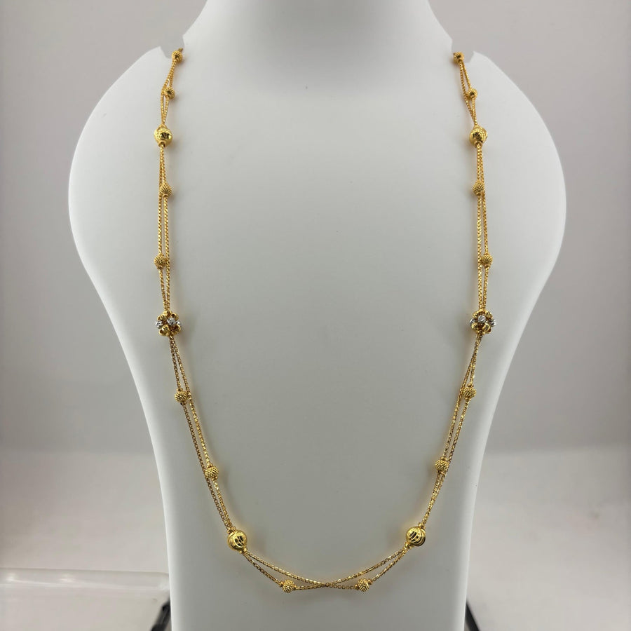 A gold necklace with small bead-like links and a central crisscross design, displayed on a white jewelry bust above a digital scale.