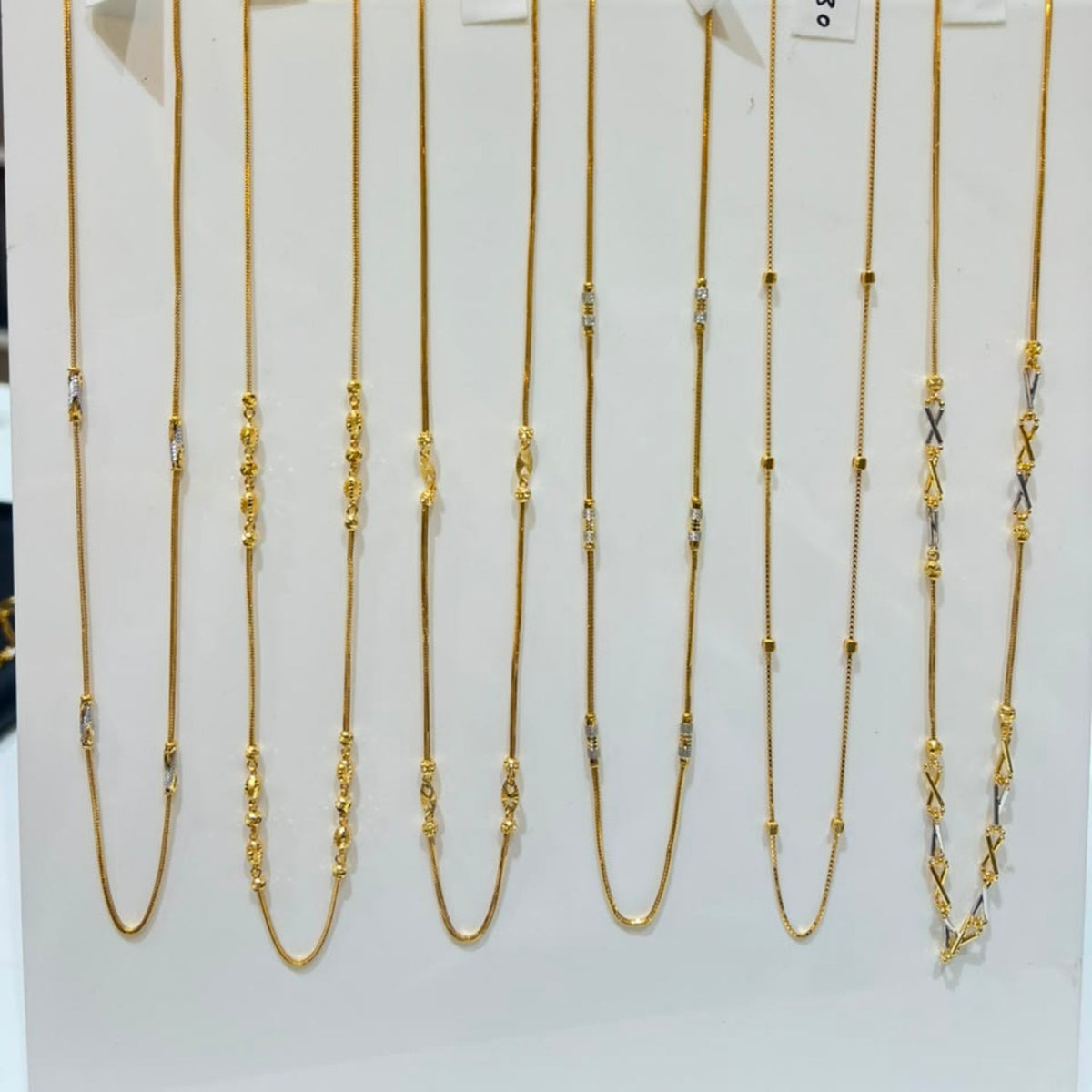 Nine gold-toned chain necklaces with various link patterns are displayed on a white vertical stand, each with a small price tag attached.