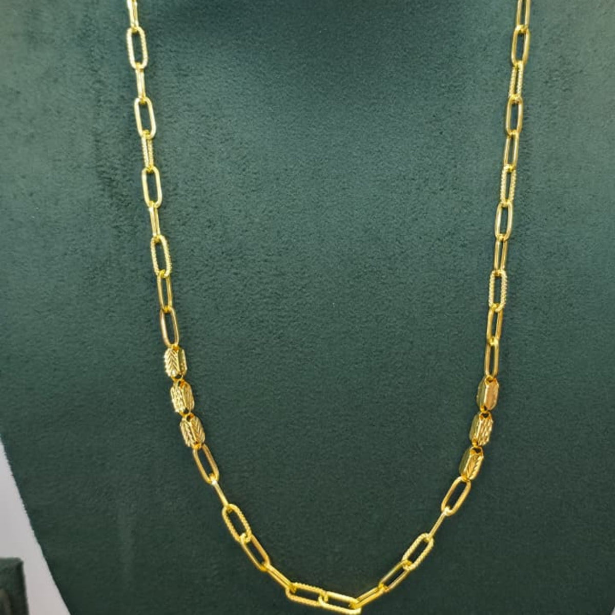 A gold chain necklace with alternating flat oval links and textured cylindrical links, displayed on a dark green velvet jewelry bust.