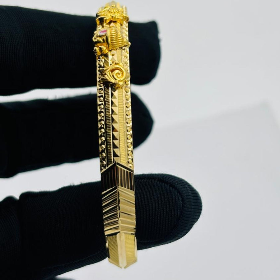 A gold-toned rectangular bar pendant or brooch with ornate sculpted details and textured geometric sections, held between fingers wearing a black glove against a plain light gray background.